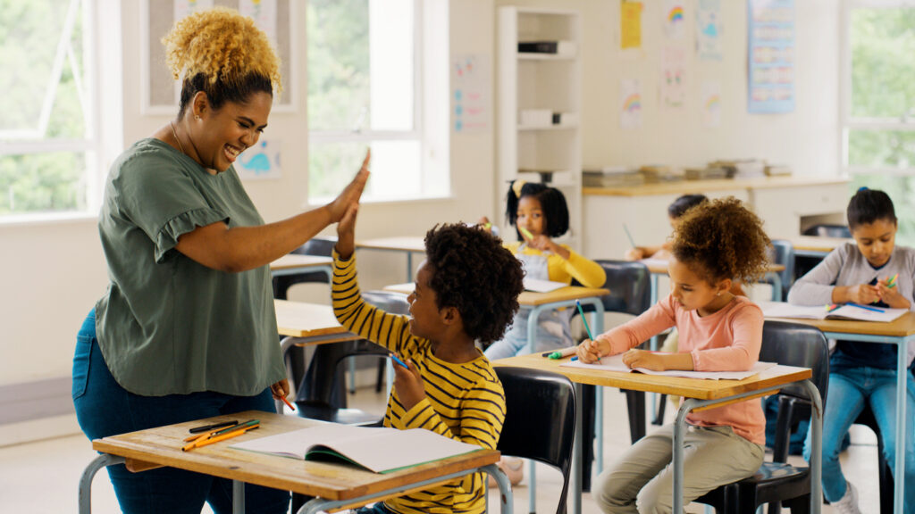 A teacher giving a high-five to a young student in a classroom while other students work at their desks.