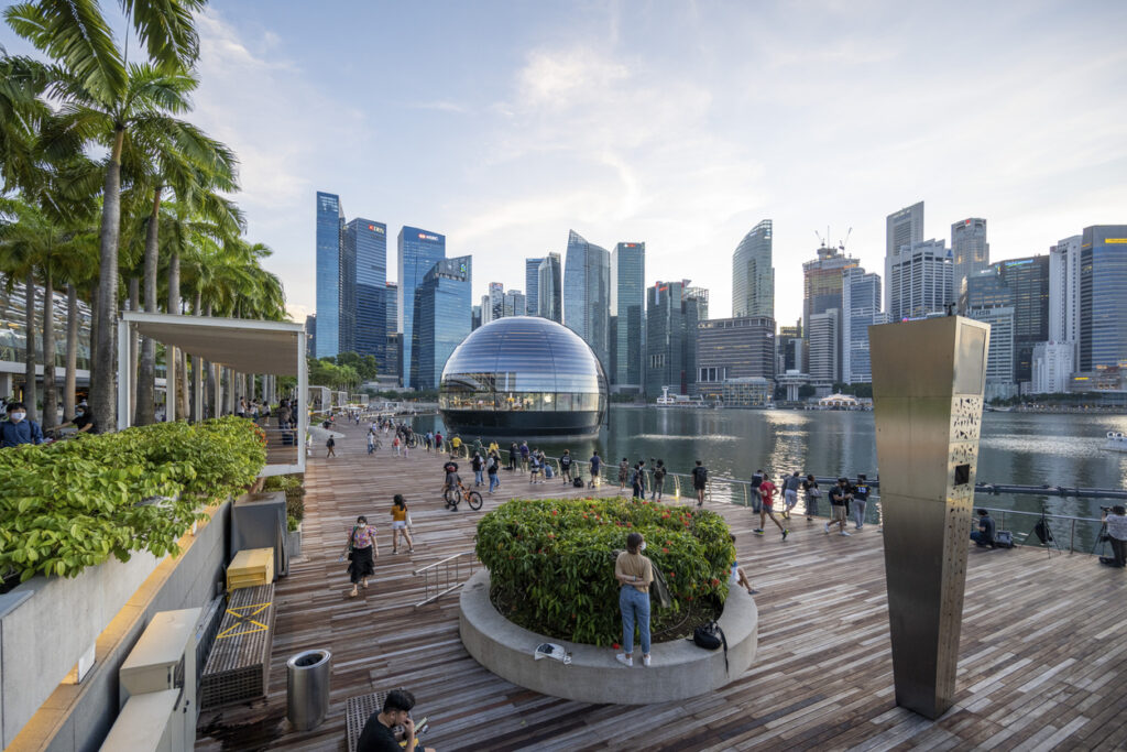 Urban waterfront promenade with pedestrians and cyclists, featuring a distinctive glass dome structure against a backdrop of modern skyscrapers.