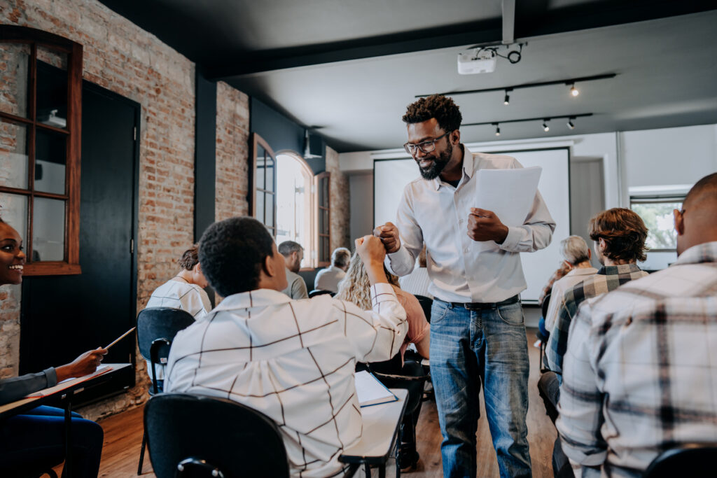 A black male presenter discussing documents with a diverse group of professionals in a modern workshop setting.