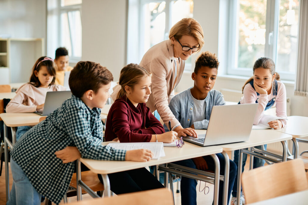 Teacher assisting a diverse group of students with a laptop in a classroom setting.