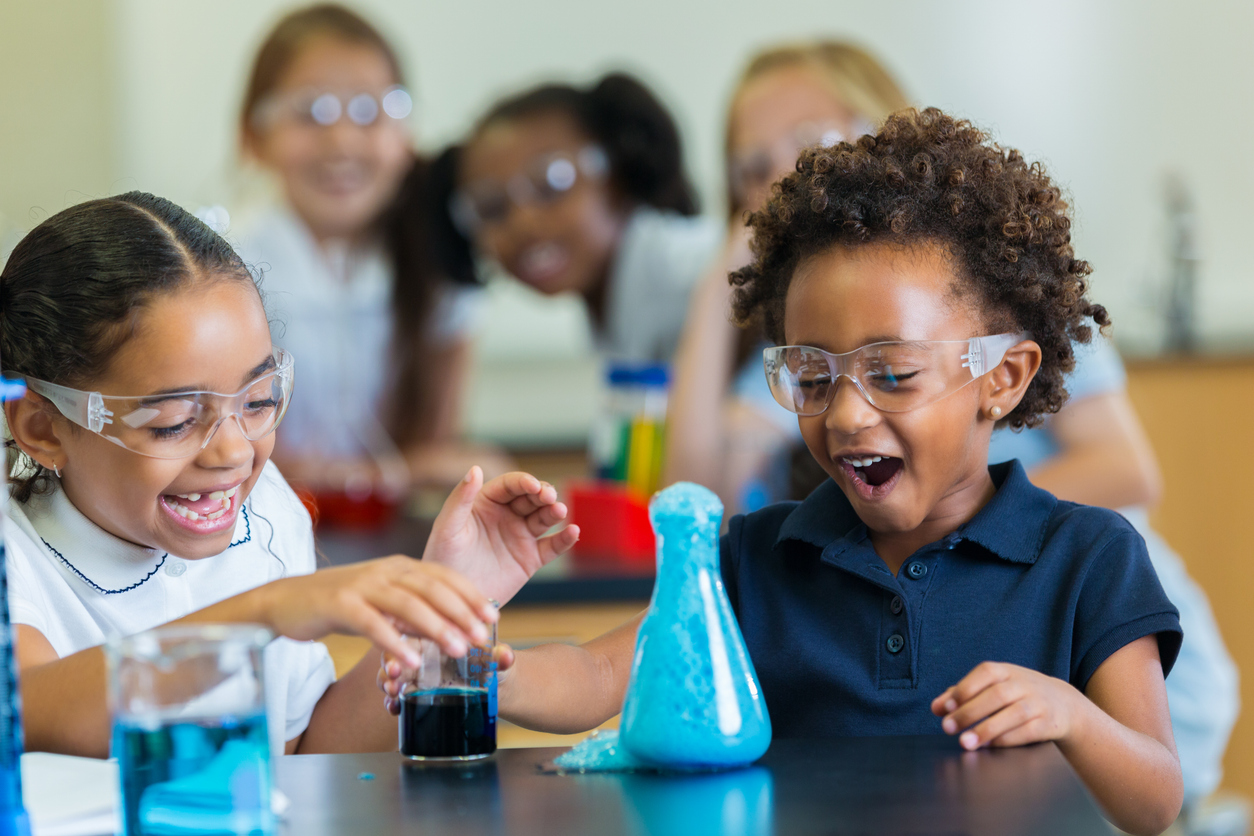 Two excited children in lab goggles watch a chemical reaction in a classroom experiment as other students look on in the background.