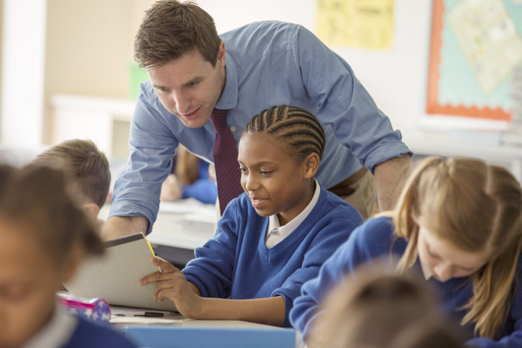 A male teacher assists a young female student in a classroom while other students work around them.