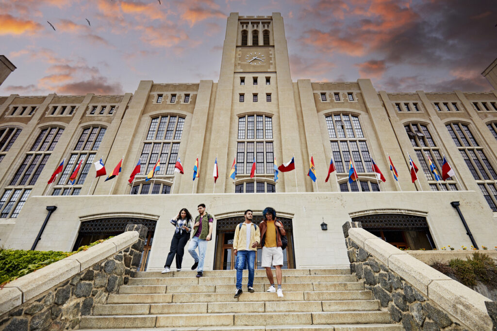 Group of students standing on stairs in front of a historic university building adorned with international flags under a dramatic sunset sky.