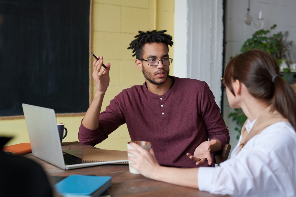 Man gesturing while having a conversation with a woman at a work table with a laptop and notebooks.