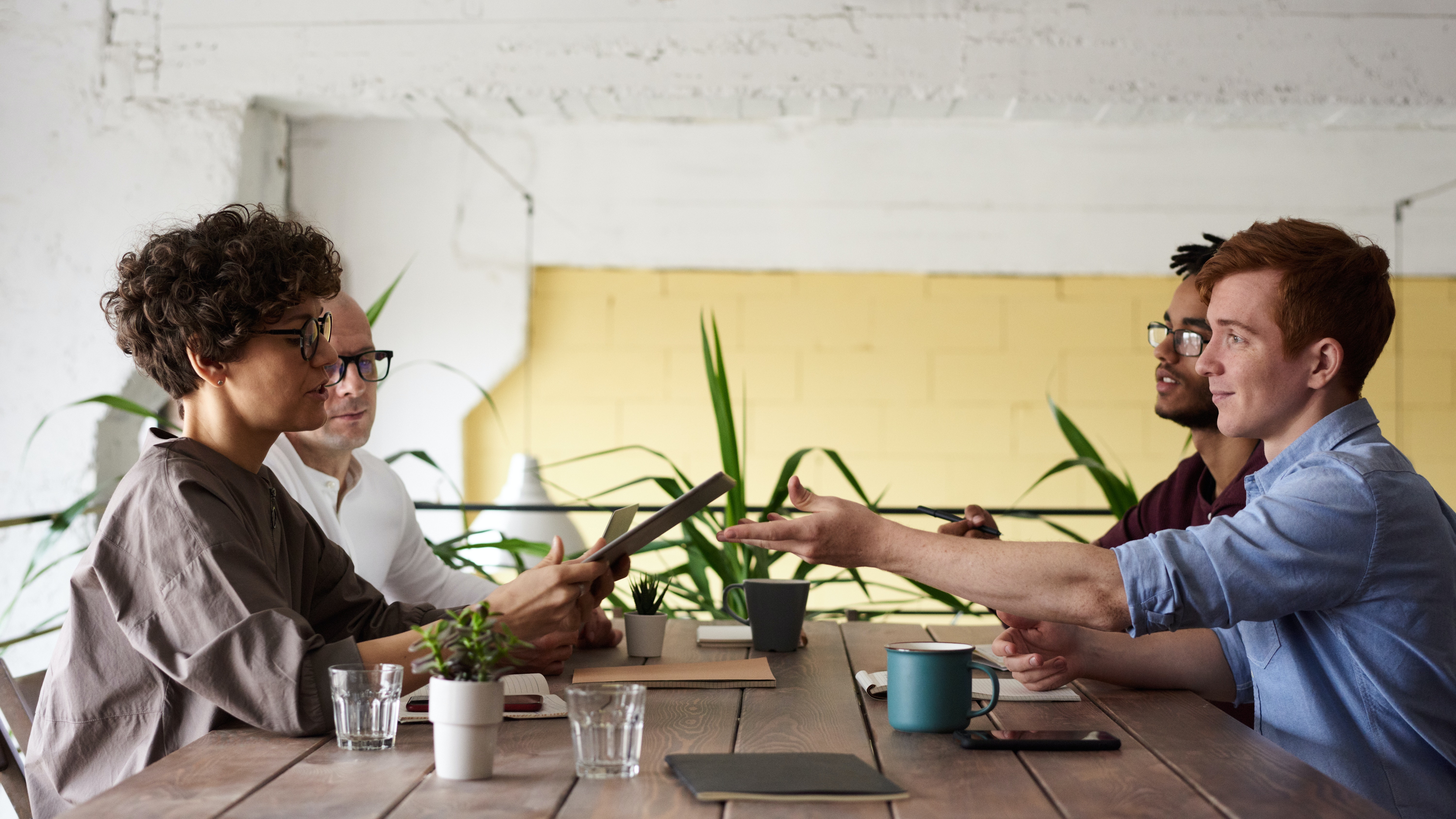 Three individuals engaged in a collaborative discussion at a meeting table, with one person presenting the budgeting plan on a tablet.