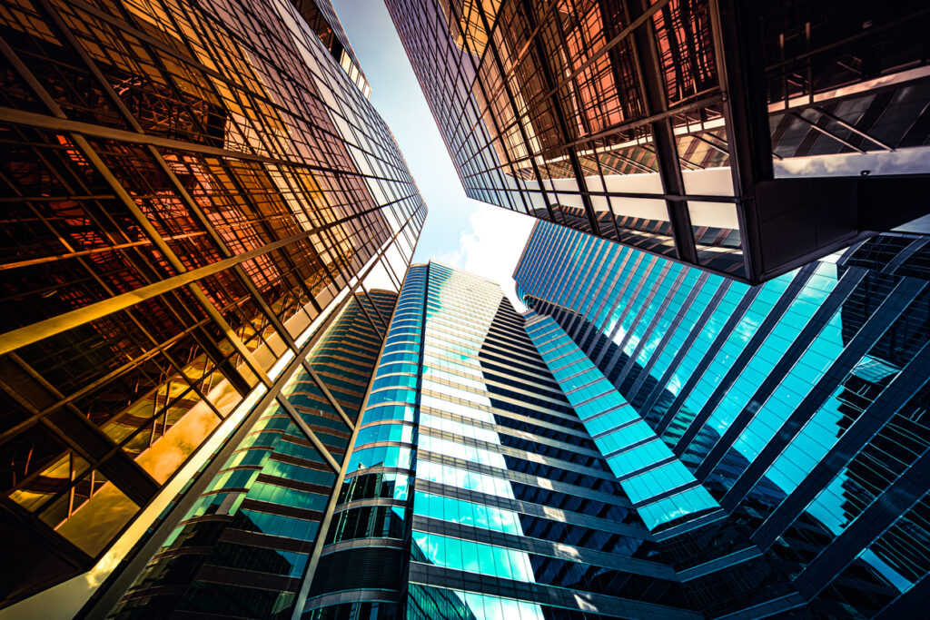 Looking up at a converging cluster of tall, modern skyscrapers with reflective glass facades against a clear blue sky.