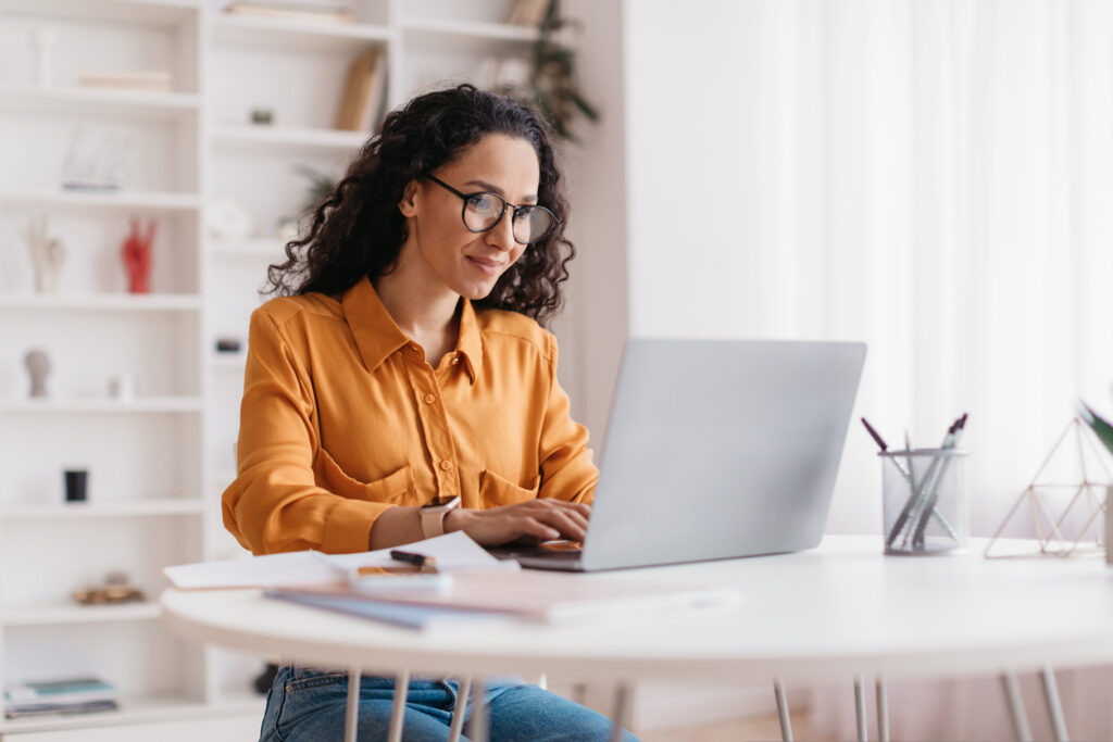 A woman with curly hair and glasses, wearing a yellow blouse, works on a laptop in a bright, modern office.