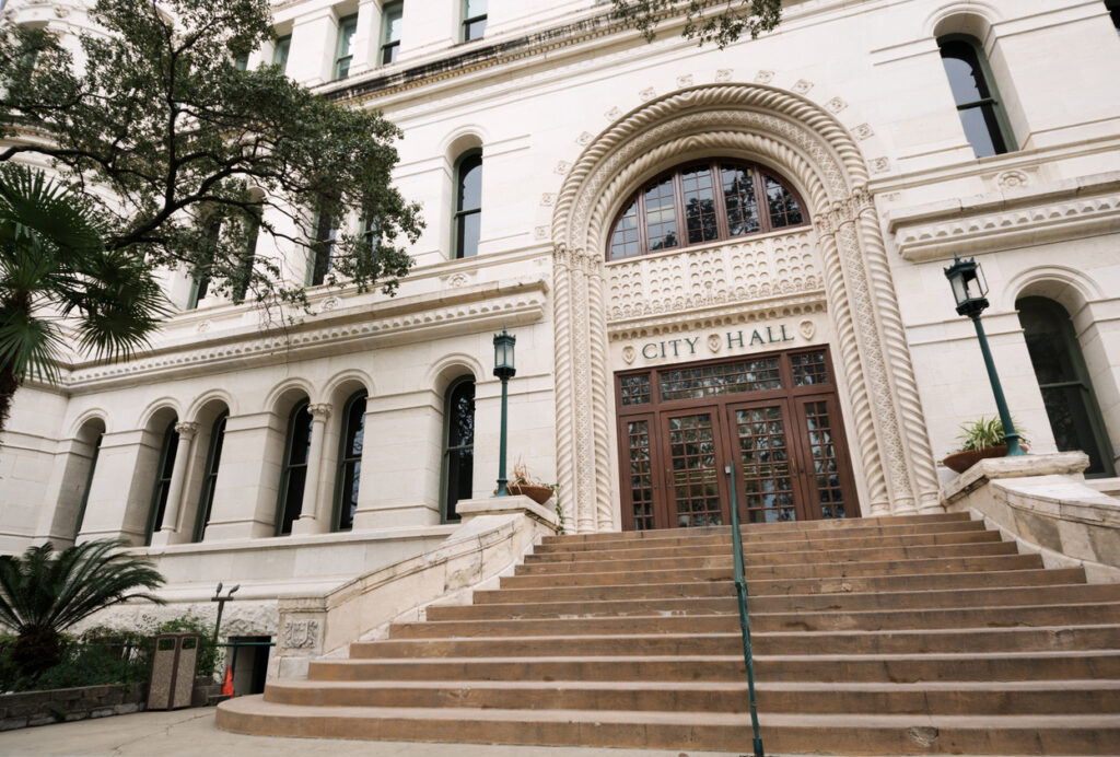 Exterior view of a grand city hall building with a large arched entryway and steps leading to a pair of bronze doors.