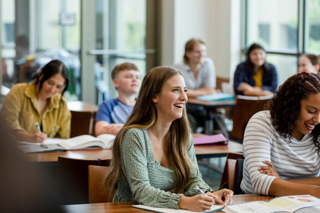 Students engaged in a classroom setting, some listening and smiling, while others are reading or taking notes.