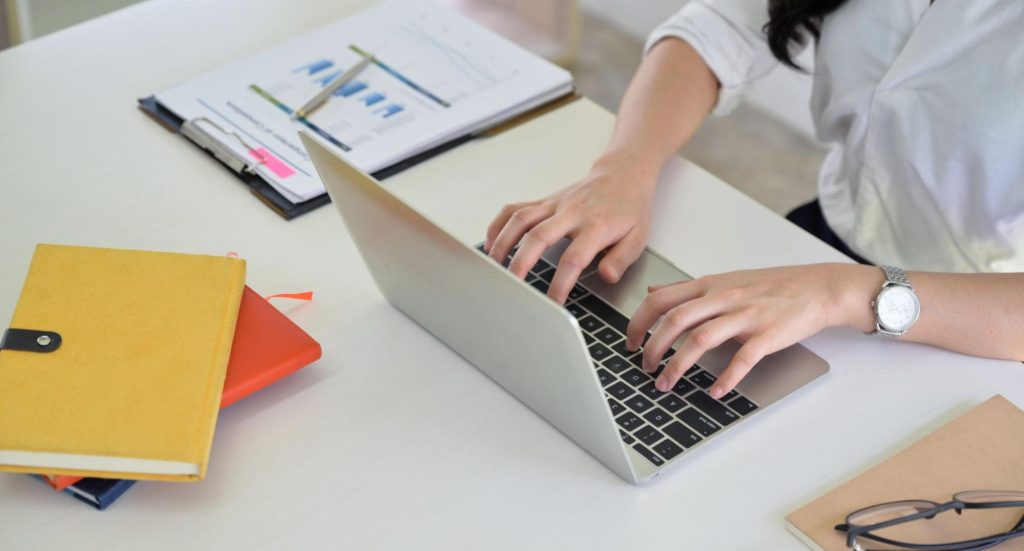 Person working on a laptop at a desk with stationery and eyeglasses nearby.