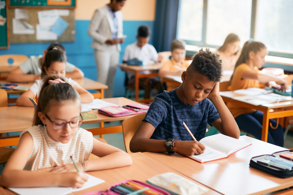 Diverse group of students focused on writing in notebooks during a classroom session with teachers supervising in the background.