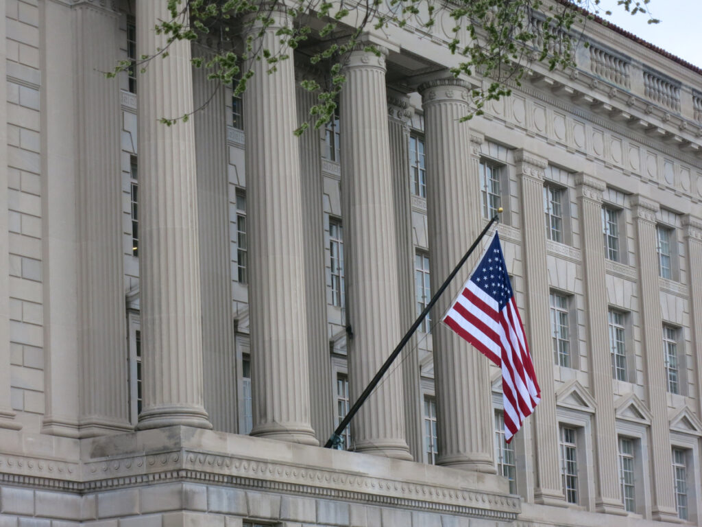 The american flag flies in front of a classical building with corinthian columns and spring foliage.