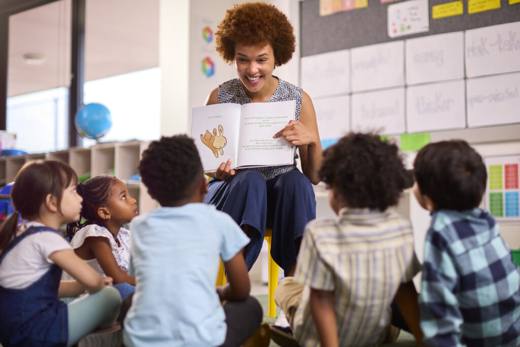 Teacher reading a book to a group of attentive young students in a classroom setting.